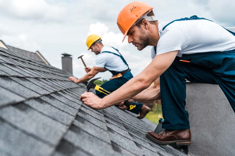two roofers wearing safety helmets repairing gray shingles on a residential roof